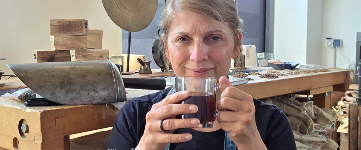 Tina mit Kaffeetasse in der Rösterei, Arbeitstisch im Hintergrund.