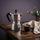 Silberner Espressokocher mit Kaffeetasse und Kaffeebohnen auf einem Holztisch vor violettem Hintergrund.