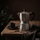 Silberner Espressokocher mit Kaffeetasse auf einem rustikalen Holztisch in dunkler, gemütlicher Atmosphäre.