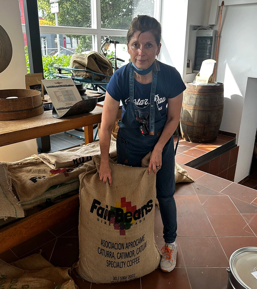 „Kaffeerösterin Tina in Arbeitsschürze aus Jeans, hält einen 69 kg schweren Kaffeesack aus Peru mit dem bunten FairBeans-Logo – symbolisiert den direkten Handel und hochwertige Kaffeebohnentransporte.“
