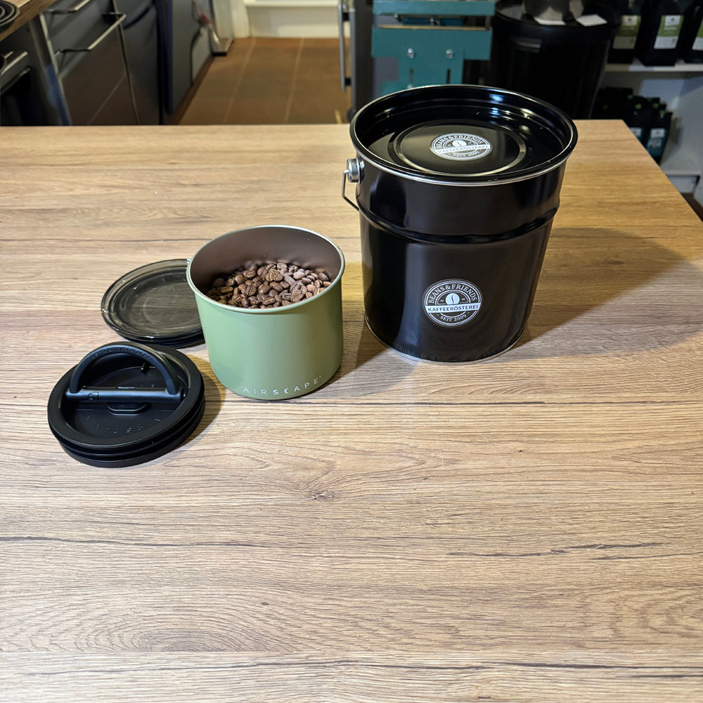 Schwarze Kaffeedose zum Nachfüllen von Beans & Friends auf einem Holztisch, daneben Kaffeezubehör.
